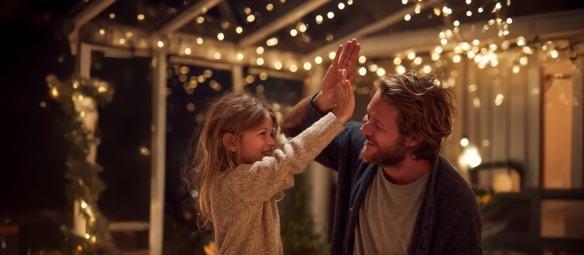 Vater und Kind klatschen sich lächelnd ab unter warmen Lichtern – Symbol für Lebensfreude, Geborgenheit und verlässliche Energie von GEW Wilhelmshaven.